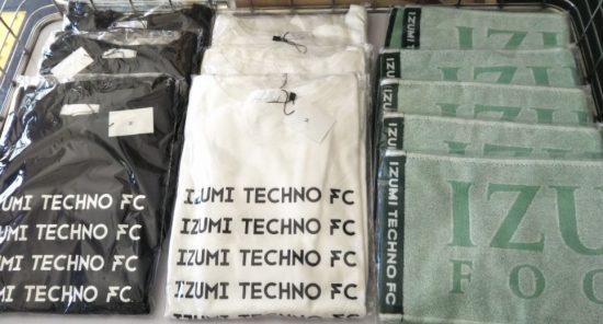 IZUMI TECHNO FCと書かれた黒と白の2種類のTシャツと緑色のタオルが展示されている写真